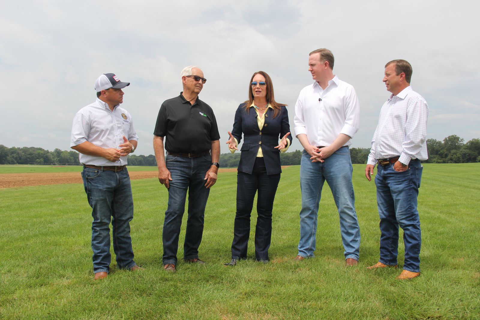 Women wearing a denim suit and yellow shirt  stands in center of four men talking. Shortest man standing on far left wears baseball cap, white shirt and jeans. Man of equal height as woman wears black polo shirt and jeans and black sunglasses. Tallest man on right of woman wears white shirt and jeans and a slightly shorter man wears white shirt and jeans. Group is standing on a green field of turf grass.