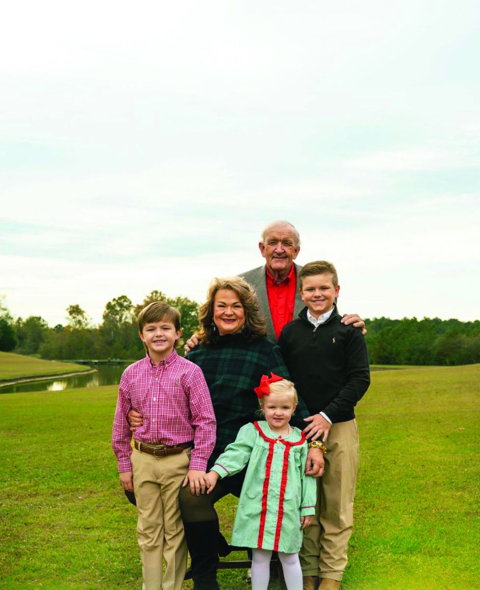 Smiling grandparents stand with their three grandchildren - two boys and 1 little girl - in a field with pond behind them