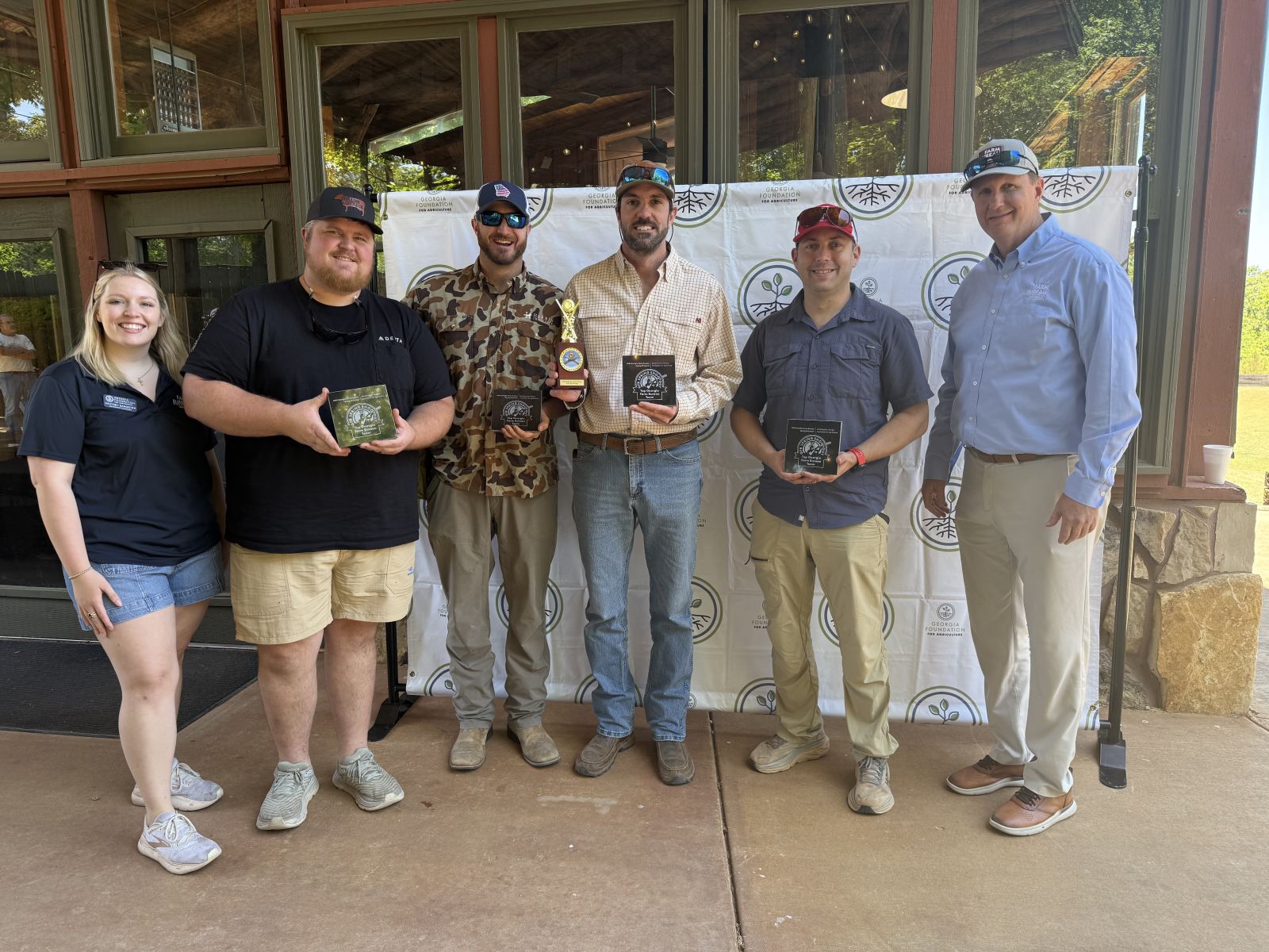 Four men dressed casually stand in center holding small square granite plaques they are accepting from young lady on far left and tall man on far right.