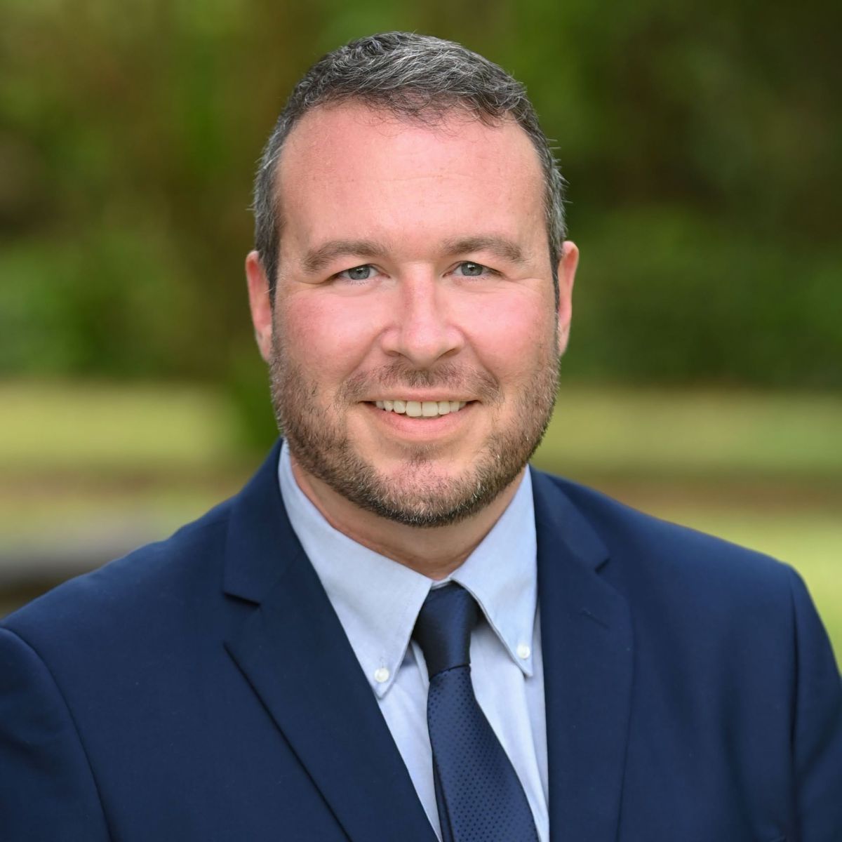 Photo of smiling white male with short black hair and closely shaven beard wearing a light blue business shirt, dark blue tie and navy sports coat.
