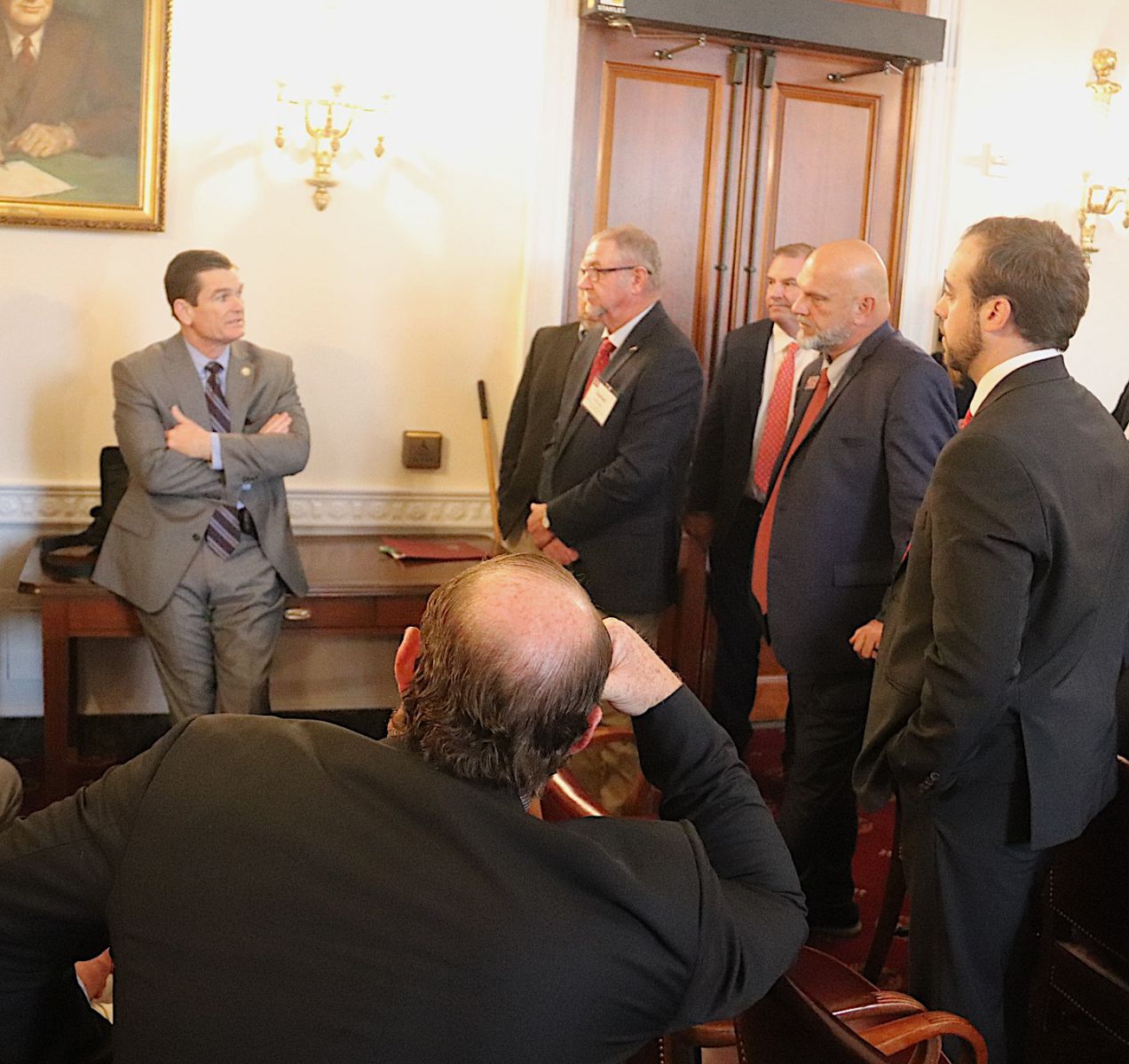 Group of six men in suits - five standing one sitting - talk to man in suit standing against a desk .