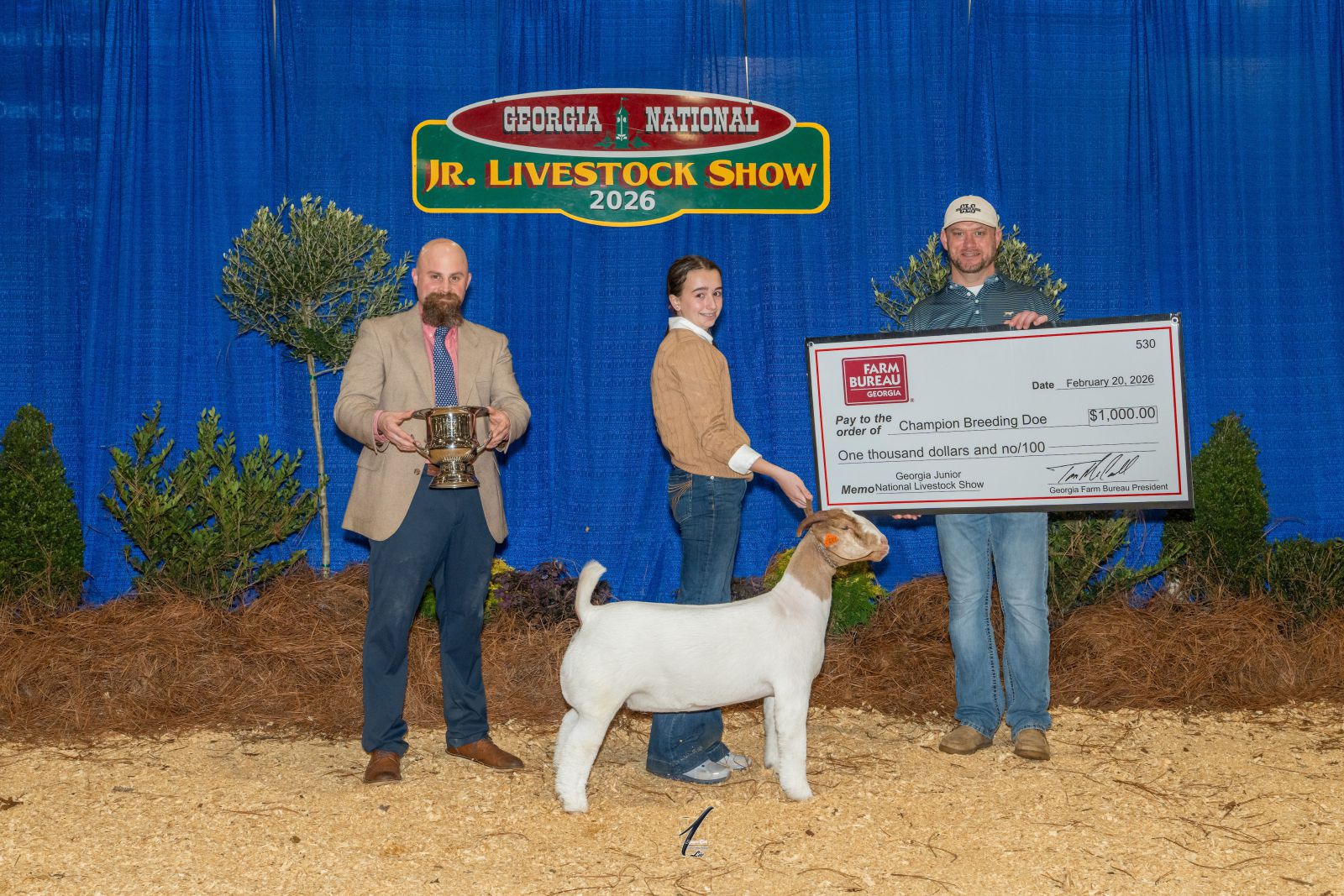 teenage girl shows goat while accepting award trophy from show judge and prize check from award sponsor.