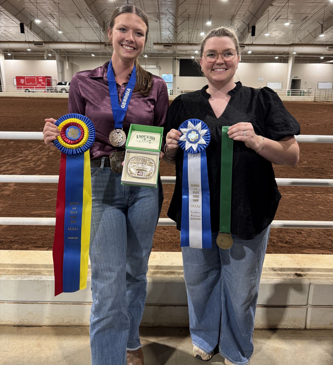 Tall teenage girl wearing silky purple long sleeve shirt and bluejeans with a blue ribbon with 40H cloverleaf medallion around her neck holds a large championship ribbon that's blue, red and yellow. Girl is also holding a green box with a championship belt buckle in it. Standing beside girl is a shorter adult who is smiling and presenting teenage with a blue and white ribbon and a green ribbon medallion that can be worn around the neck. 