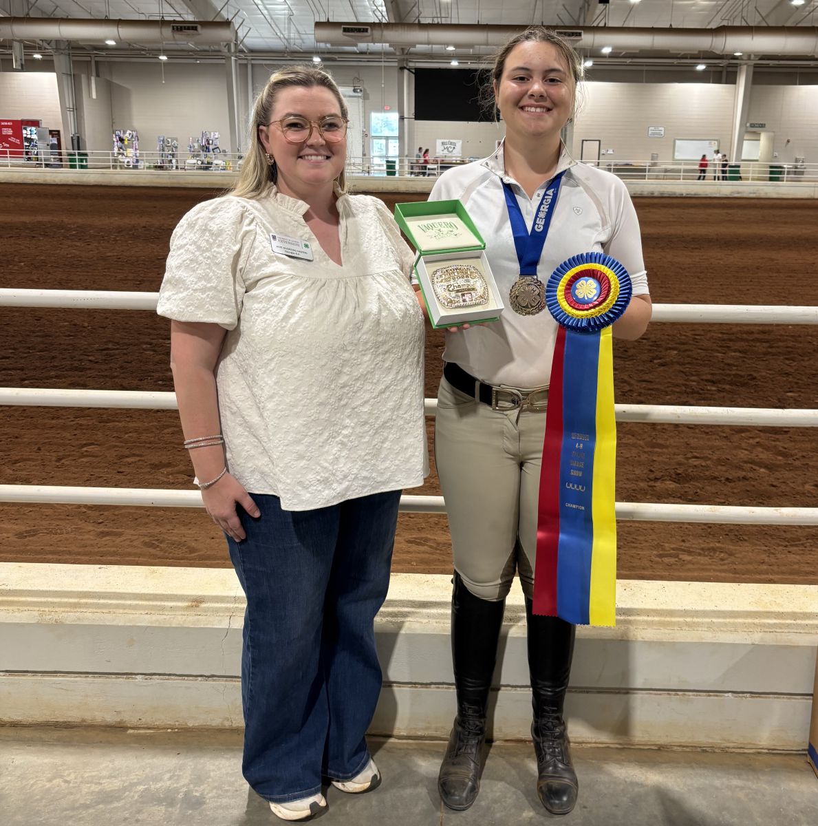 Smiling female adult stands on left congratulating a slightly taller smiling teenage girl who is wearing a white polo shirt, kakhi riding pants and English riding boots. Teenage girl is wearing a blue ribbon medallion around her neck and holding a green box with a championship belt buckle in one hand and a championship ribbon that's blue, red and yellow in the other hand.