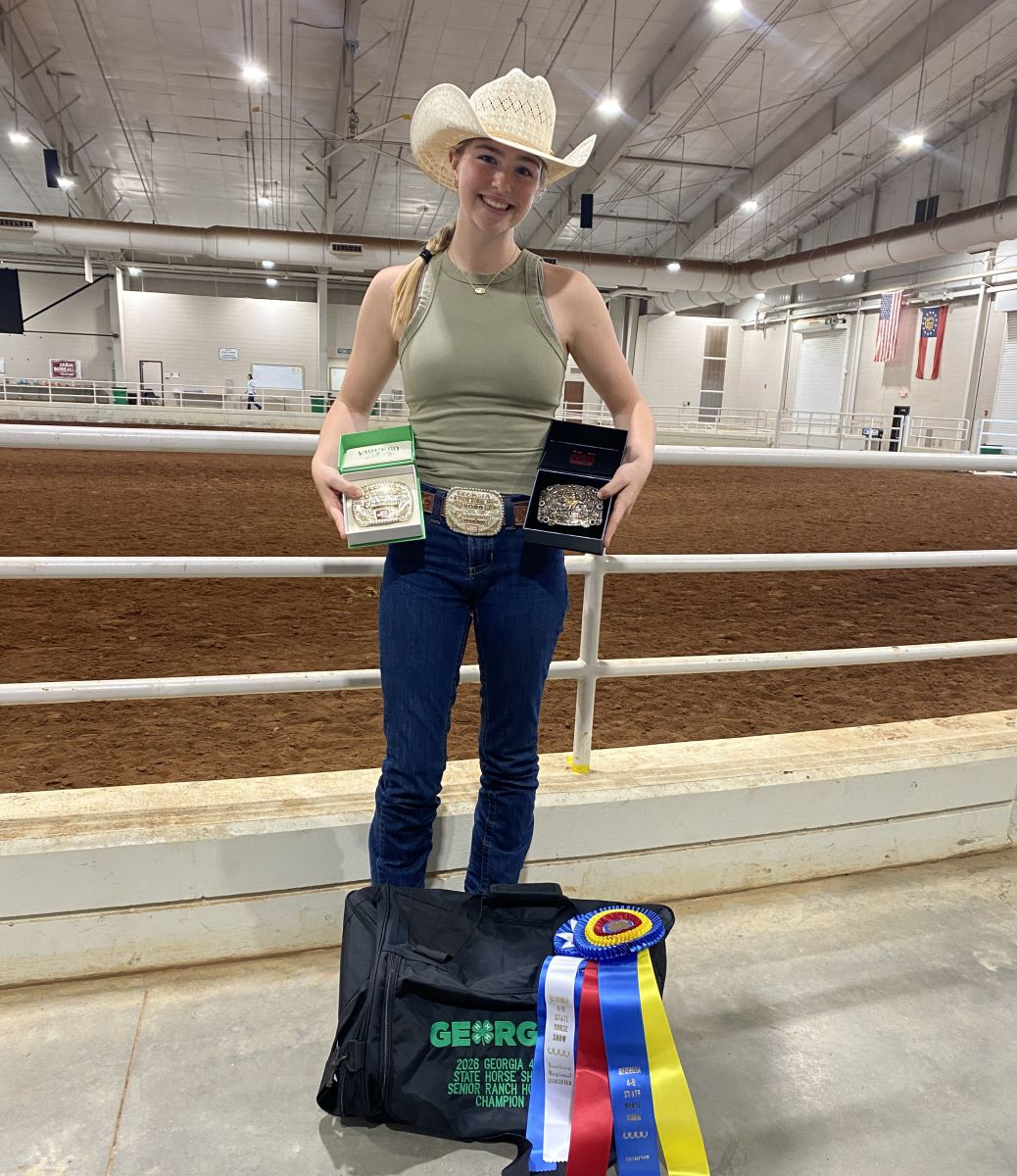 Teenage girl wearing beige cowboy hat, sage green tank top, and jeans with a belt buckle holds a box with a championship belt buckle in each hand. At her feet sits a black duffle bag with Georgia 4-H Horse Show in green lettering. Blue, white, red and yellow ribbons lay on the corner of the duffel bag.
