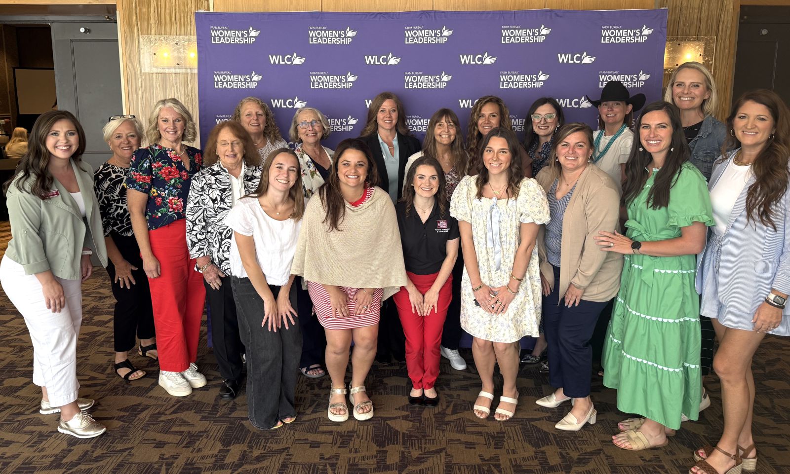 Group of 19 women varying ages stand smiling in front of a purple backdrop