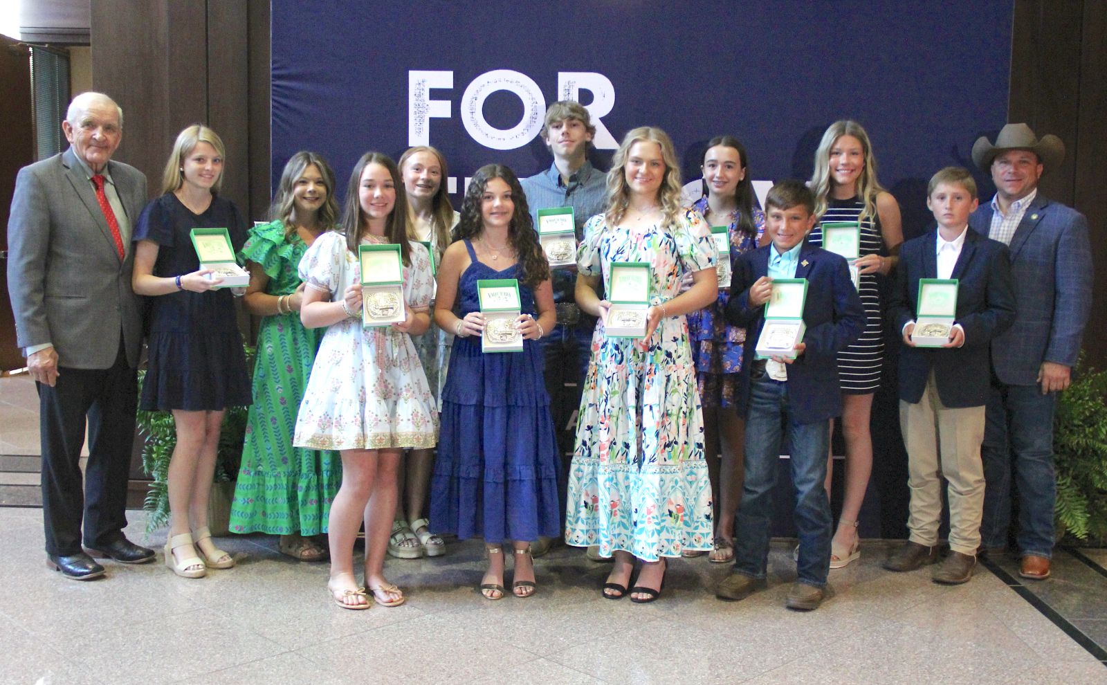 Group of 11 preteens and teenagers wearing dresses and suits hold belt buckles they won. Group stands between tall man on far left and a man wearing a cowboy hat on far right .