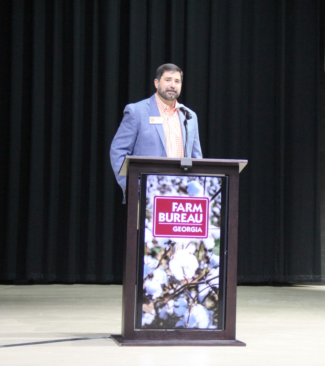 Man with black hair and beard wearing a light blue sports coat and orange gingham shirt stands behind a podium speaking into microphone. Podium has a digital photo of a cotton crop with white cotton bolls ready to harvest with a burgandy rectangle that has Farm Bureau Georgia written in white laid over a portion of the photo.