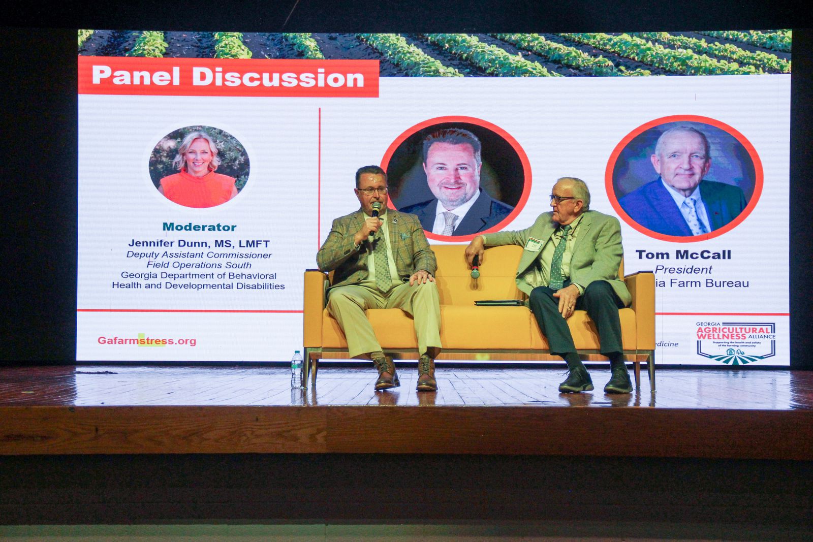 Two men wearing suits sit on a yellow couch on a stage participating in a panel discussion. Man on left holds microphone.