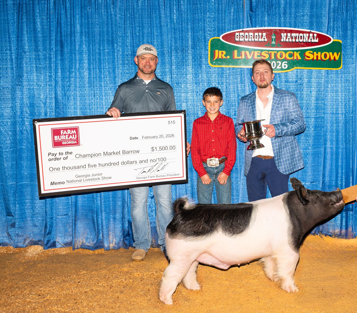 Preteen boy stands behind his prize pig and between prize sponsor presenting him a check and show judge presenting him a trophy.