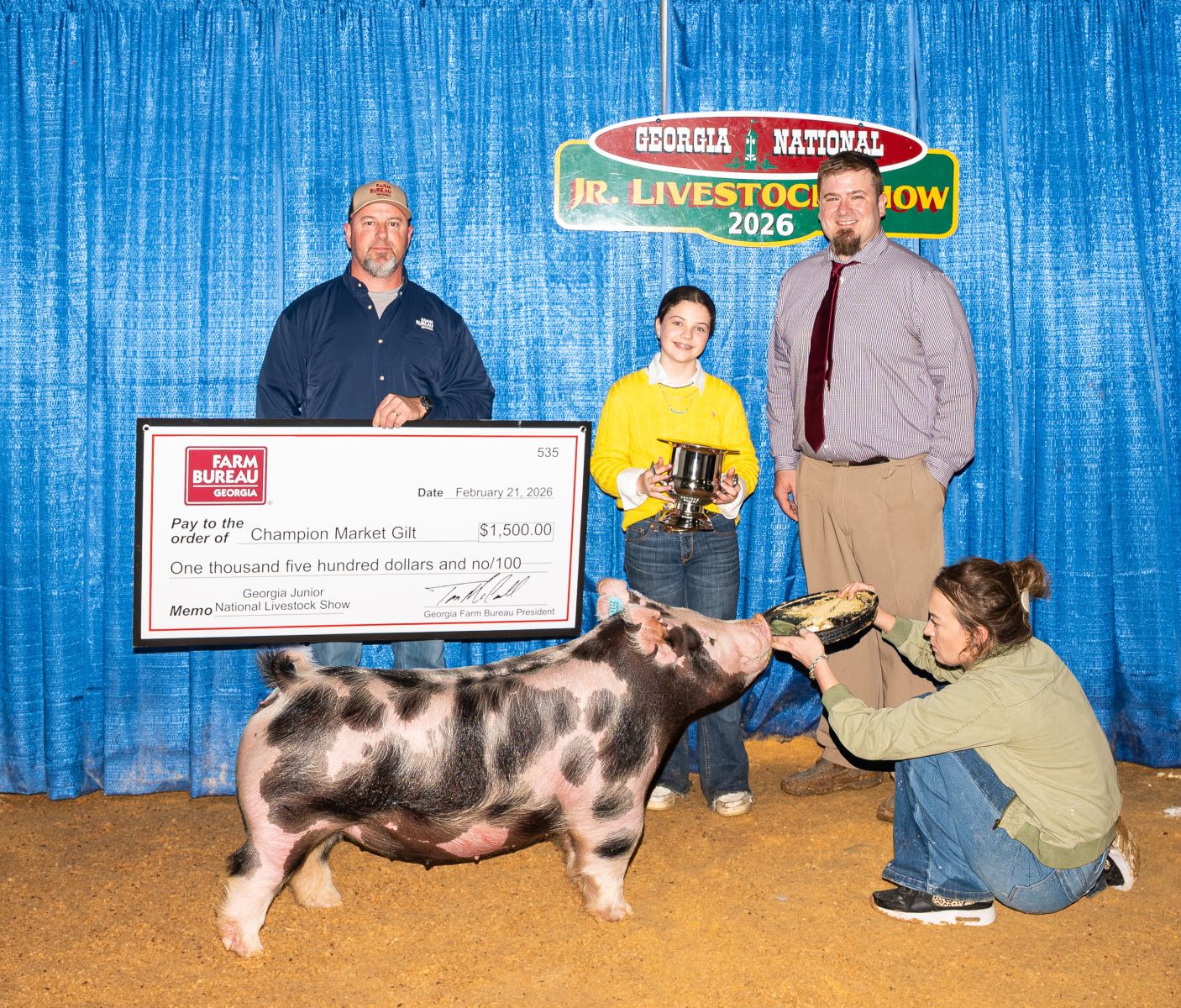 Preteen girl in yellow sweater stands behind her winning pig holding a trophy. She stands between the award sponsor on left presenting her with prize check and judge on right offering congratulations. Unknown lady kneels in front of pig holding its food bowl to keep pig in place for photo. 