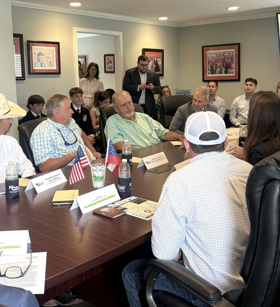 Crowded room of people. Six people are shown sitting at a table. A man wearing a green shirt is talking while others listen. People are seated in chairs against walls and three people can be seen standing.