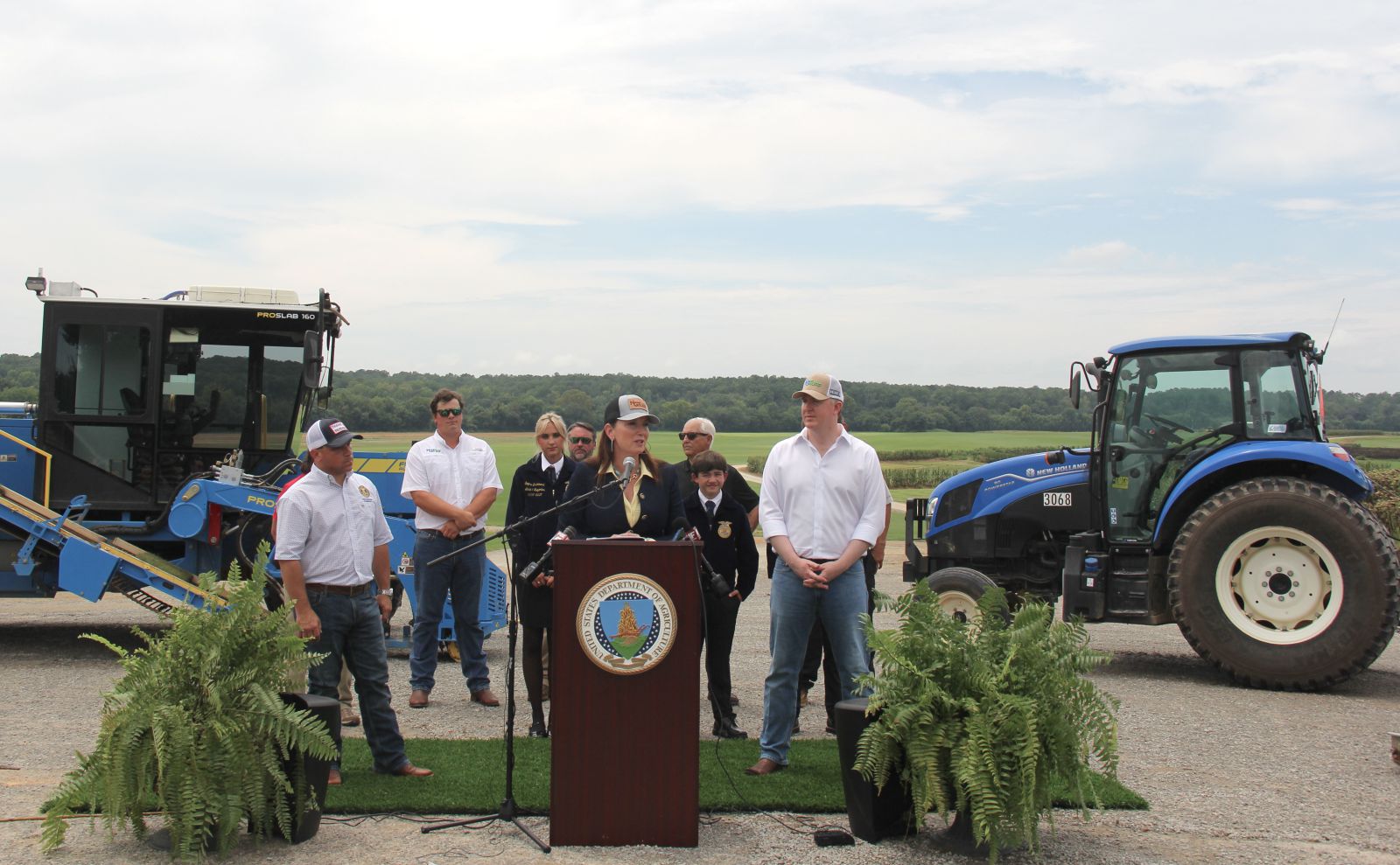 Group of eight people stand between a blue sod harvester machine and a blue tractor. Group is standing on a green artificial grass carpet with two large ferns in front of them on either side of a podium that has the USDA logo on it. Women in denim jacket and yellow shirt wearing a baseball hat stands at podium talking.