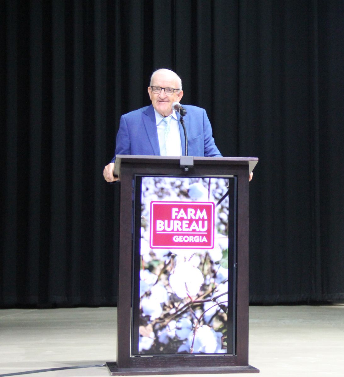 Smiling man with white hair earing black glasses and a blue sports coat, light blue shirt and tie speaks into microphone at podium that has a digital photo of a cotton plant with white cotton bolls ready to harvest. A burgandy rectangle with Farm Bureau Georgia in white letters covers a small portion of the cotton plant photo.