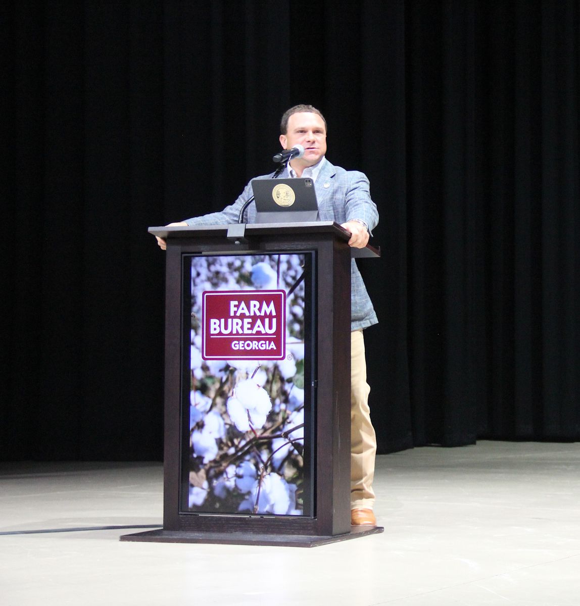 Young man with short dark brown hair wearing a light blue plaid sportscoat, light blue shirt, khaki pants with a sharp crease, and brown cowboy boots stands behind podium talking into microphone. Podium has a photo of cotton with white cotton bolls ready to be harvested that has a burgandy rectangle with Farm Bureau Georgia written in white letters over part of photo.