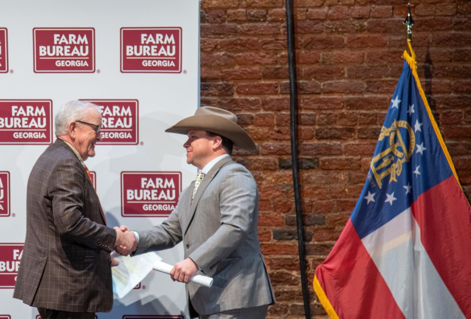 Two men in suits shake hands as man on left exits stage and man on right wearing cowboy hat walks onto stage.