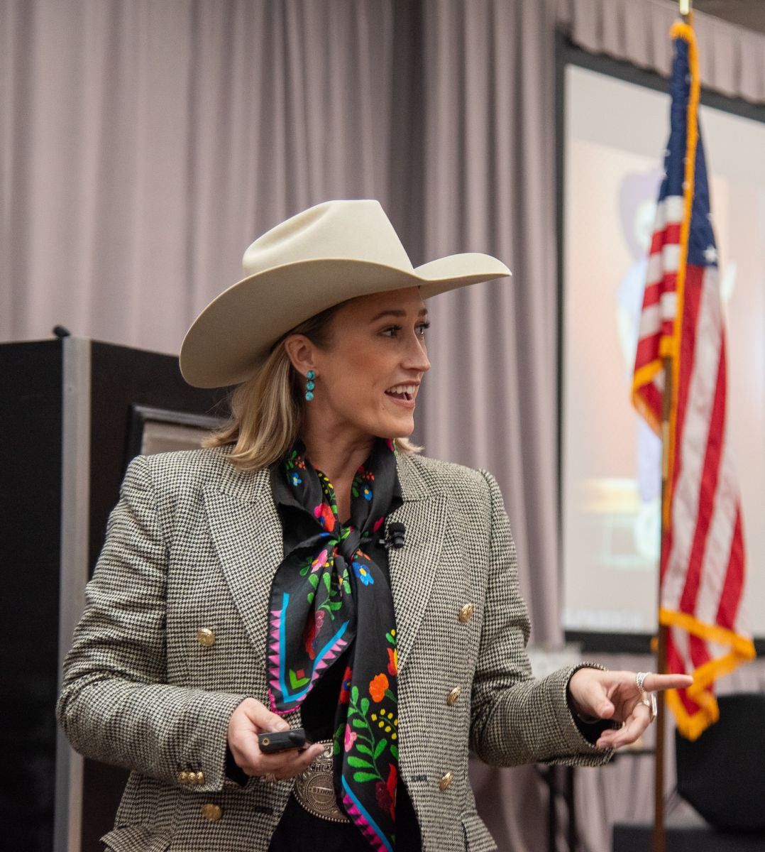 Smiling lady dressed in beige cowboy hat and suit gives speech in front of a U.S. flag