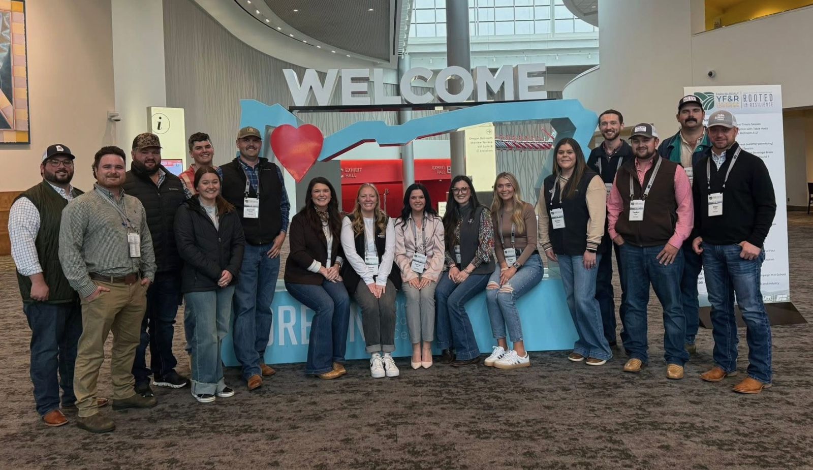 A group of 16 young people dressed in casual clothes - 9 guys and 7 ladies - stand around or sit a large Welcome to Oregon sign.