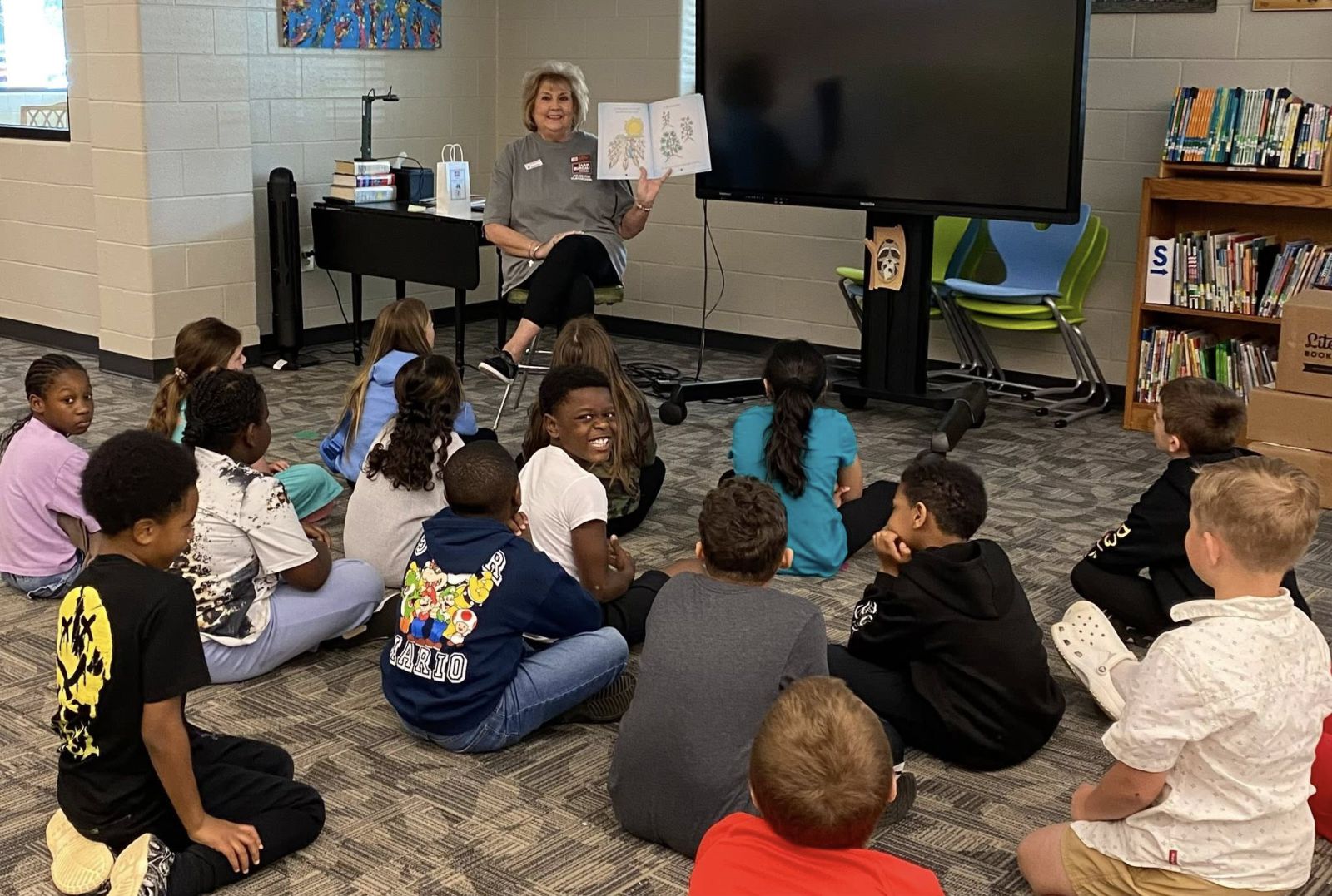 A woman sitting in a chair reading a book to a classroom full of students seated on the floor.