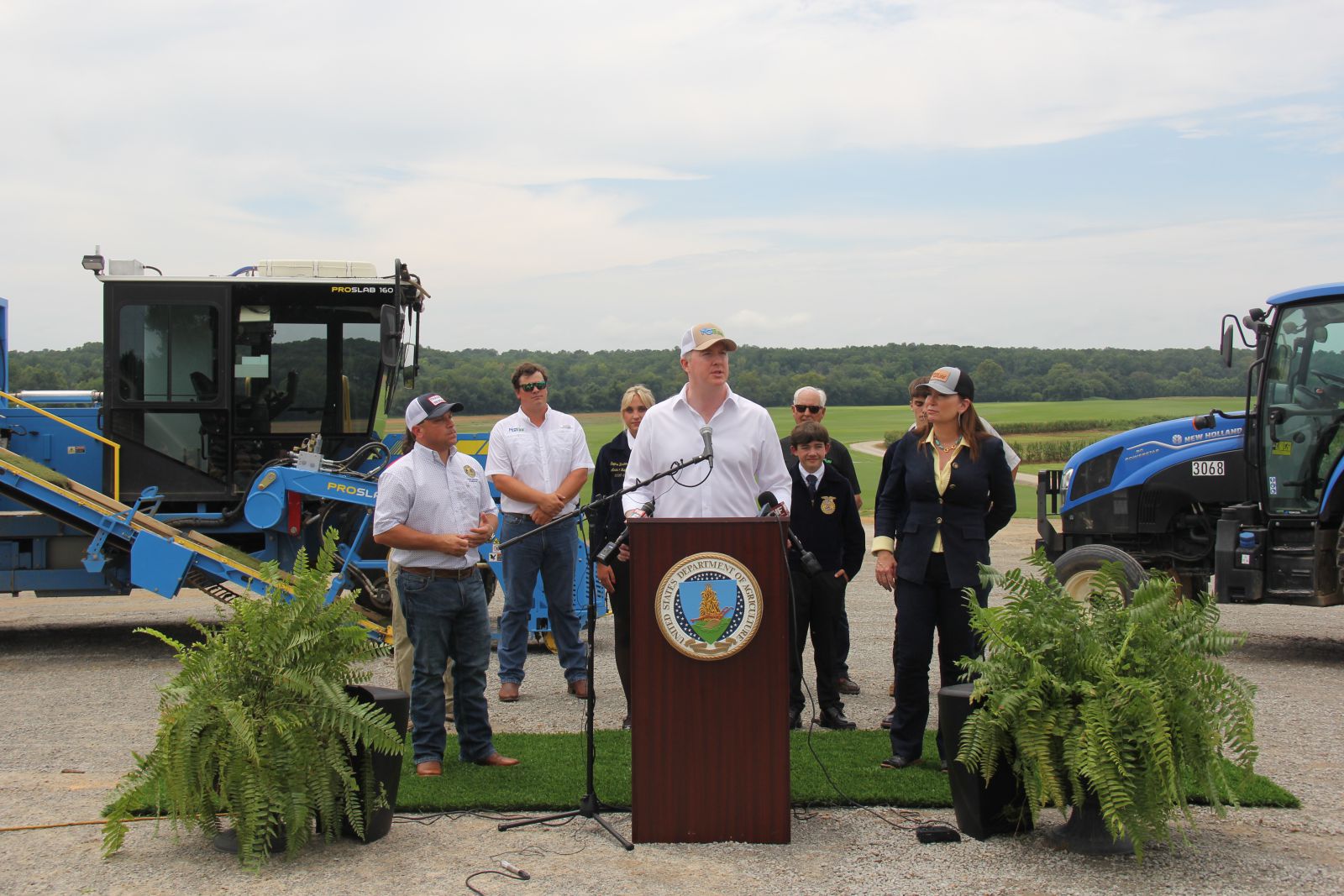Tall man wearing a khaki baseball cap and white shirt stands behind podium talking. Group of about seven people stand behind him between a blue sod harvester machine and a blue tractor. Group is standing on a green artificial grass carpet with two large ferns on either side of podium.